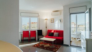 A modern living room with a red sofa, a white coffee table, and a red and beige rug. There is a dining area with red chairs and a white table. The room has large windows with a view of buildings and a balcony with white furniture.