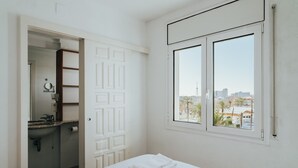 A hotel room with a bed, a window with a city view, a bathroom with a sink and mirror, and a door leading to another room.
