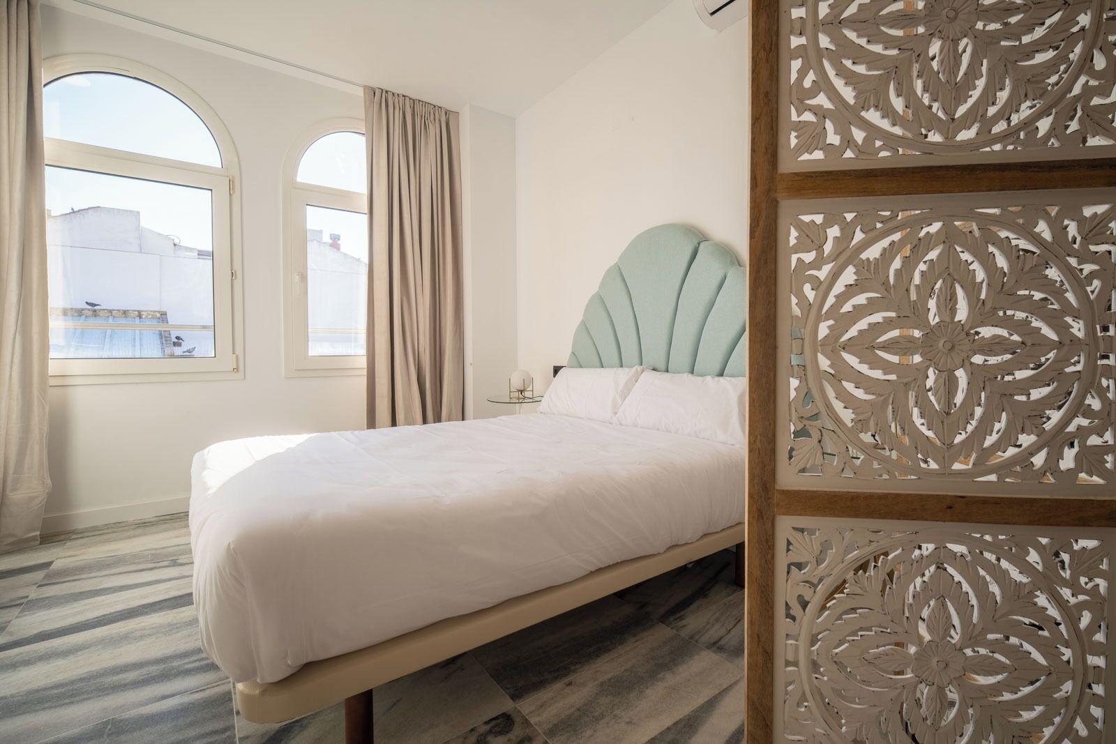 A bed with a light blue headboard, a window with a view of buildings, and a decorative panel with intricate patterns.