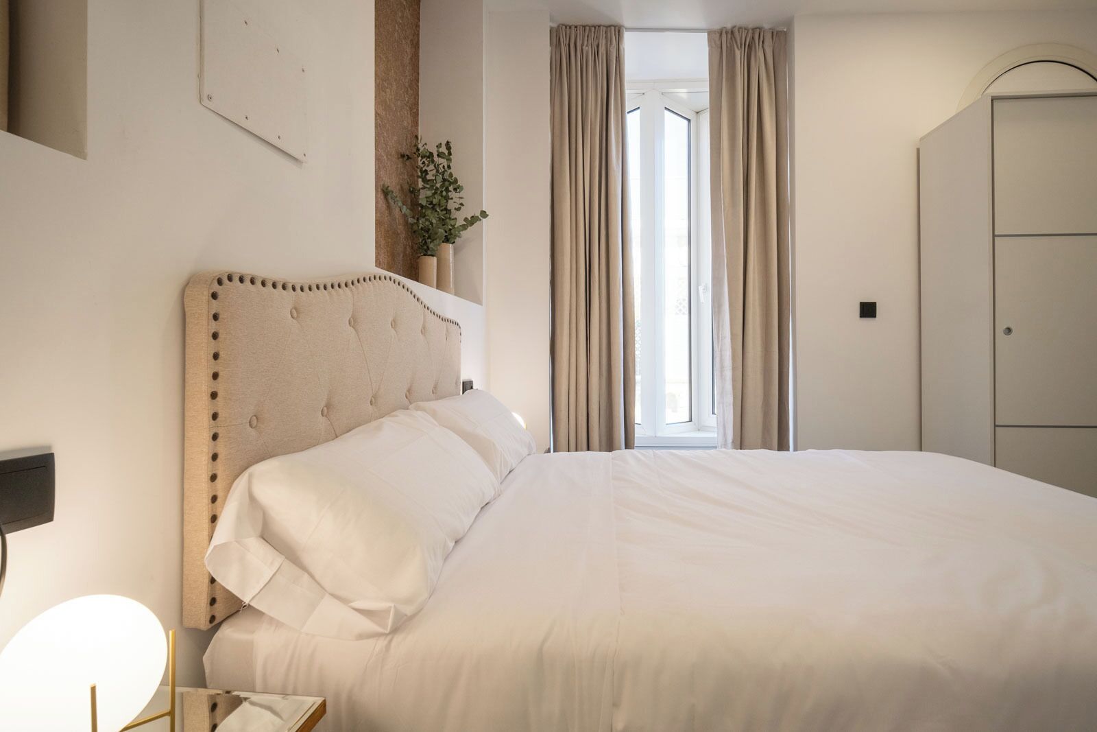 A neatly made bed with a tufted headboard in a hotel room with a large window and curtains.