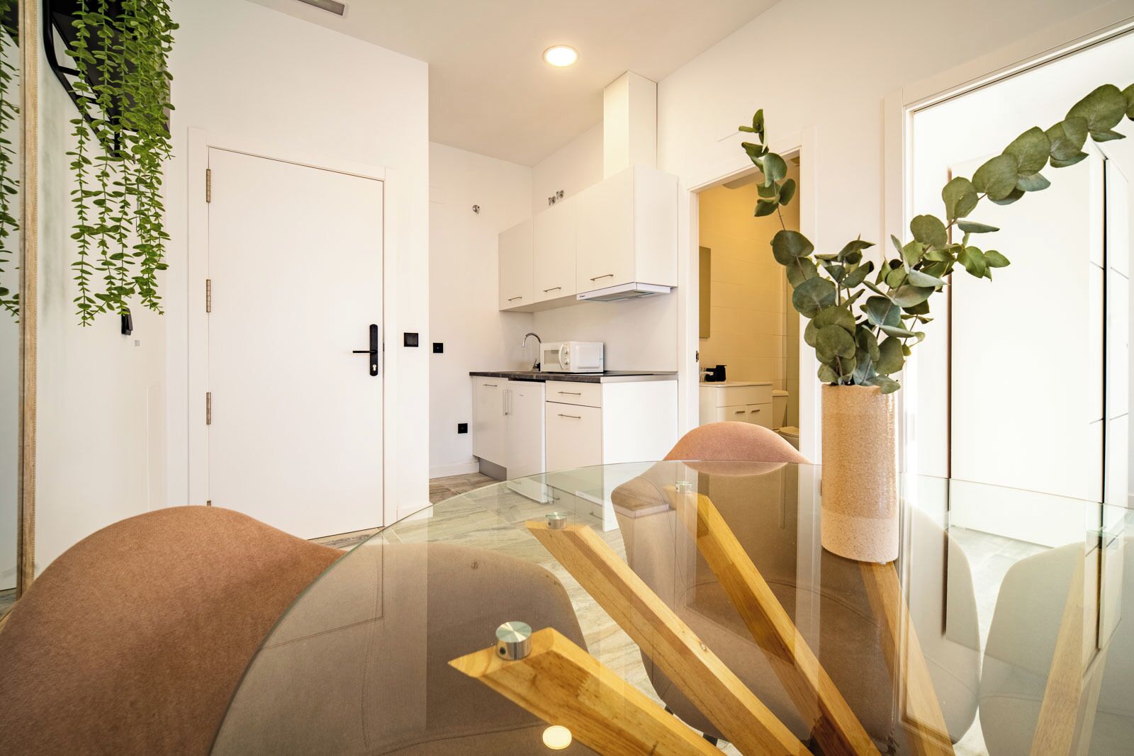 A modern kitchen with white cabinets, a microwave, and a sink. There is a glass table with a wooden top and a plant in a pot.