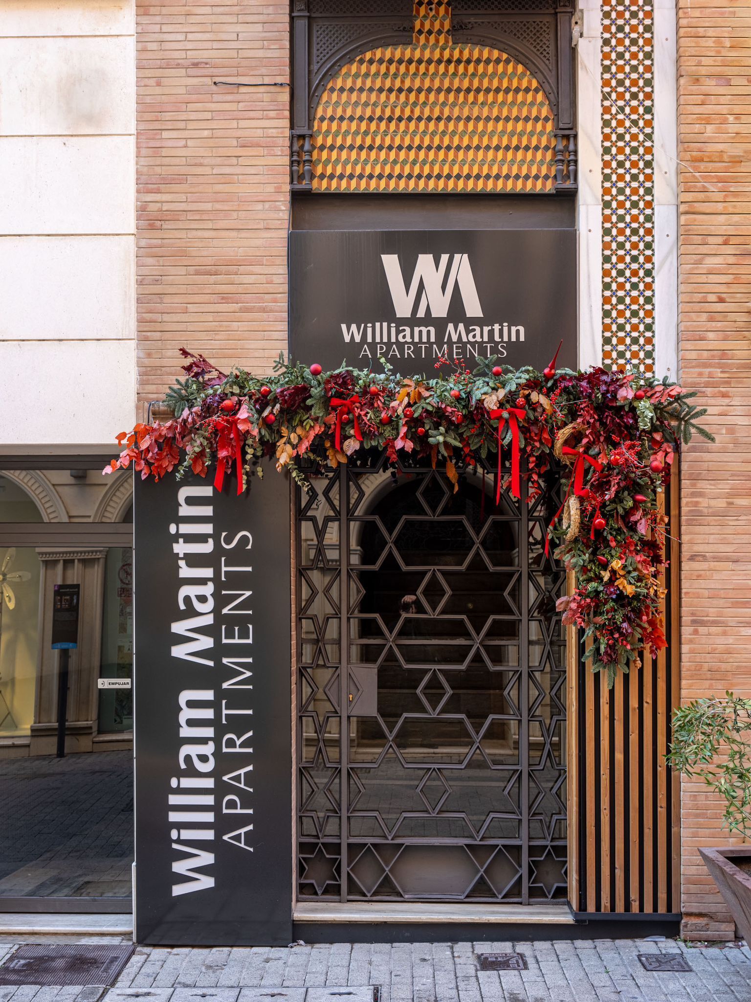 Een gebouwentree met een bordje voor William Martin Apartments, met een decoratieve krans en een geometrisch metalen hek.