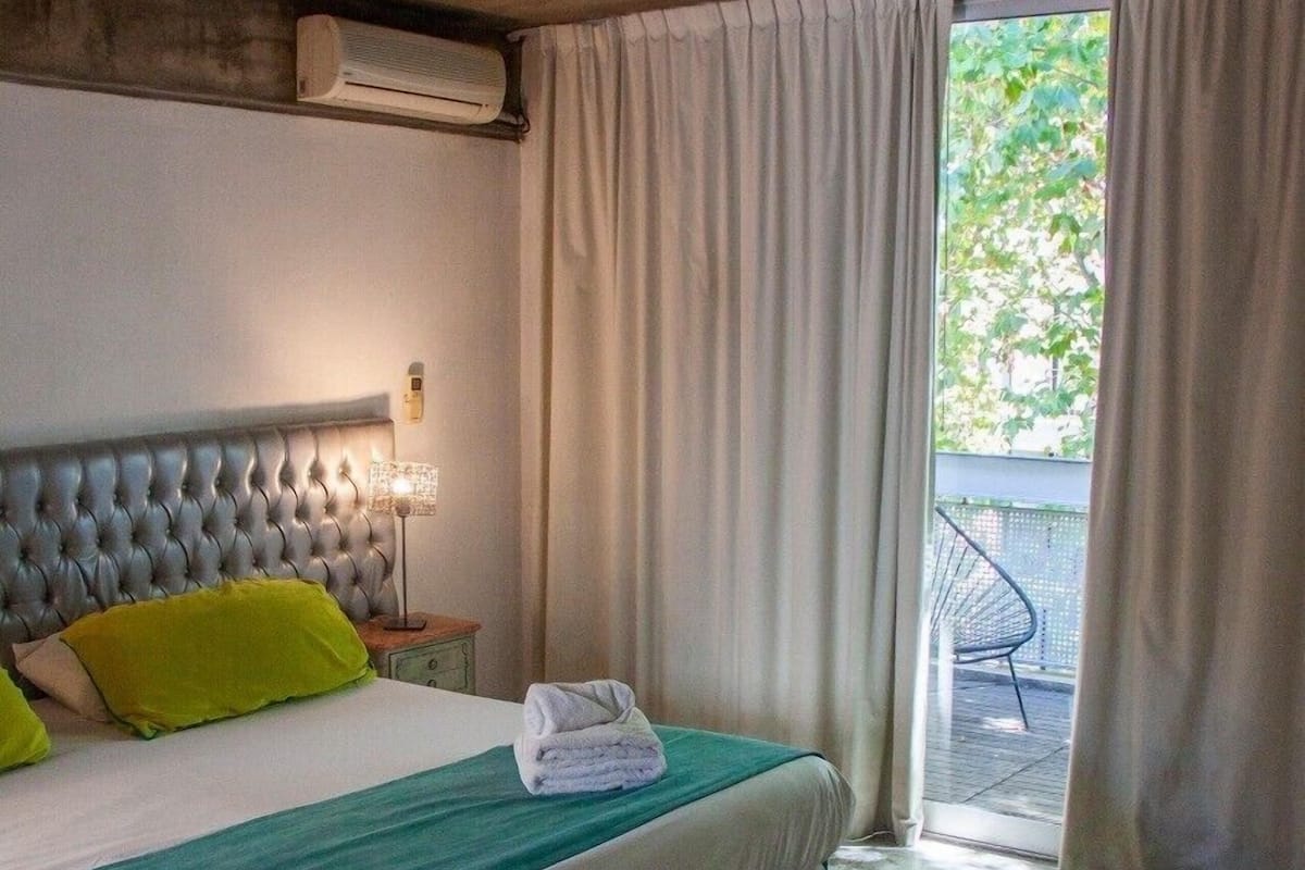 A bedroom with a large bed, a nightstand with a lamp, a window with curtains, and a view of the outdoors.