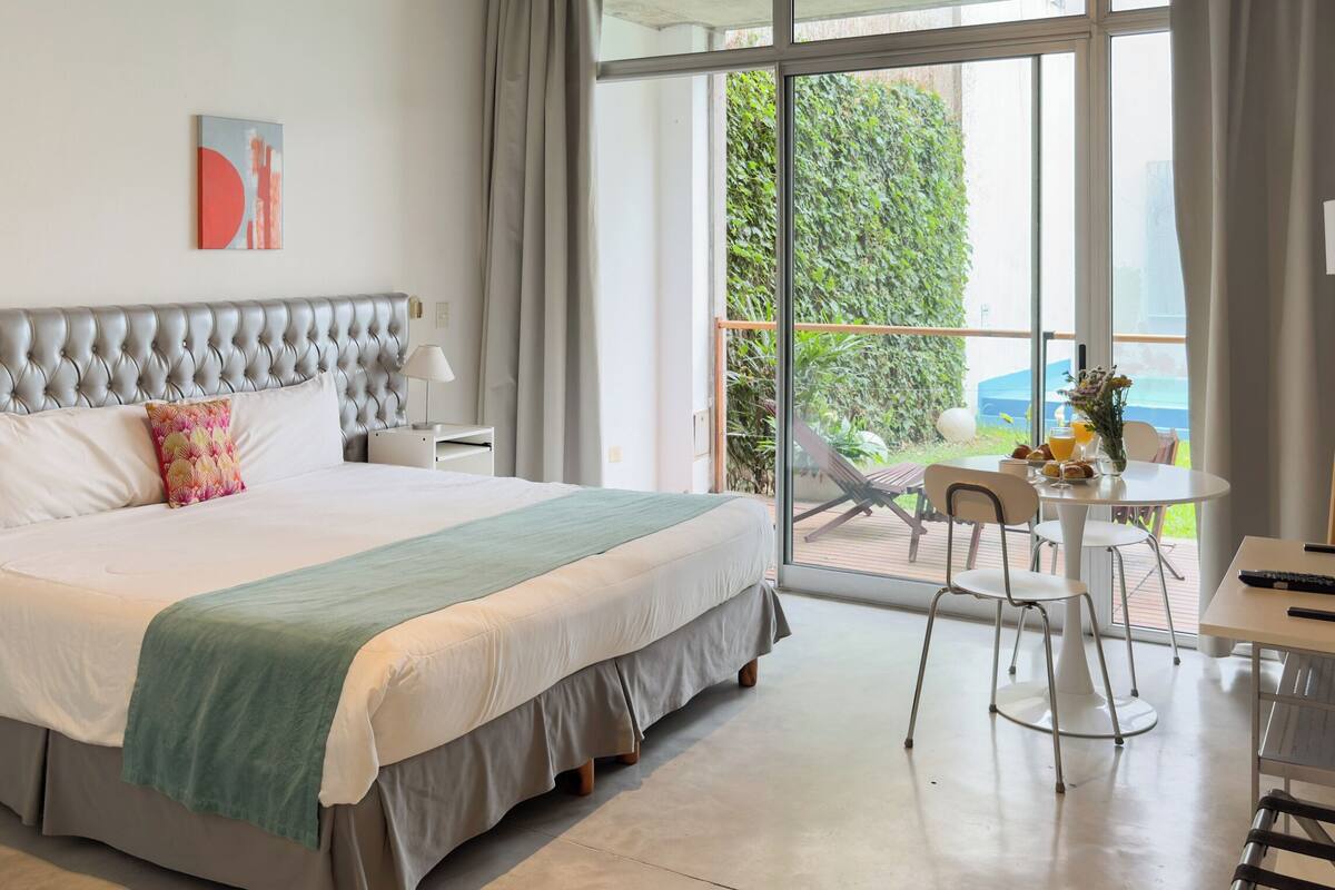 A modern hotel room with a large bed, a desk, and a balcony with a table and chairs.