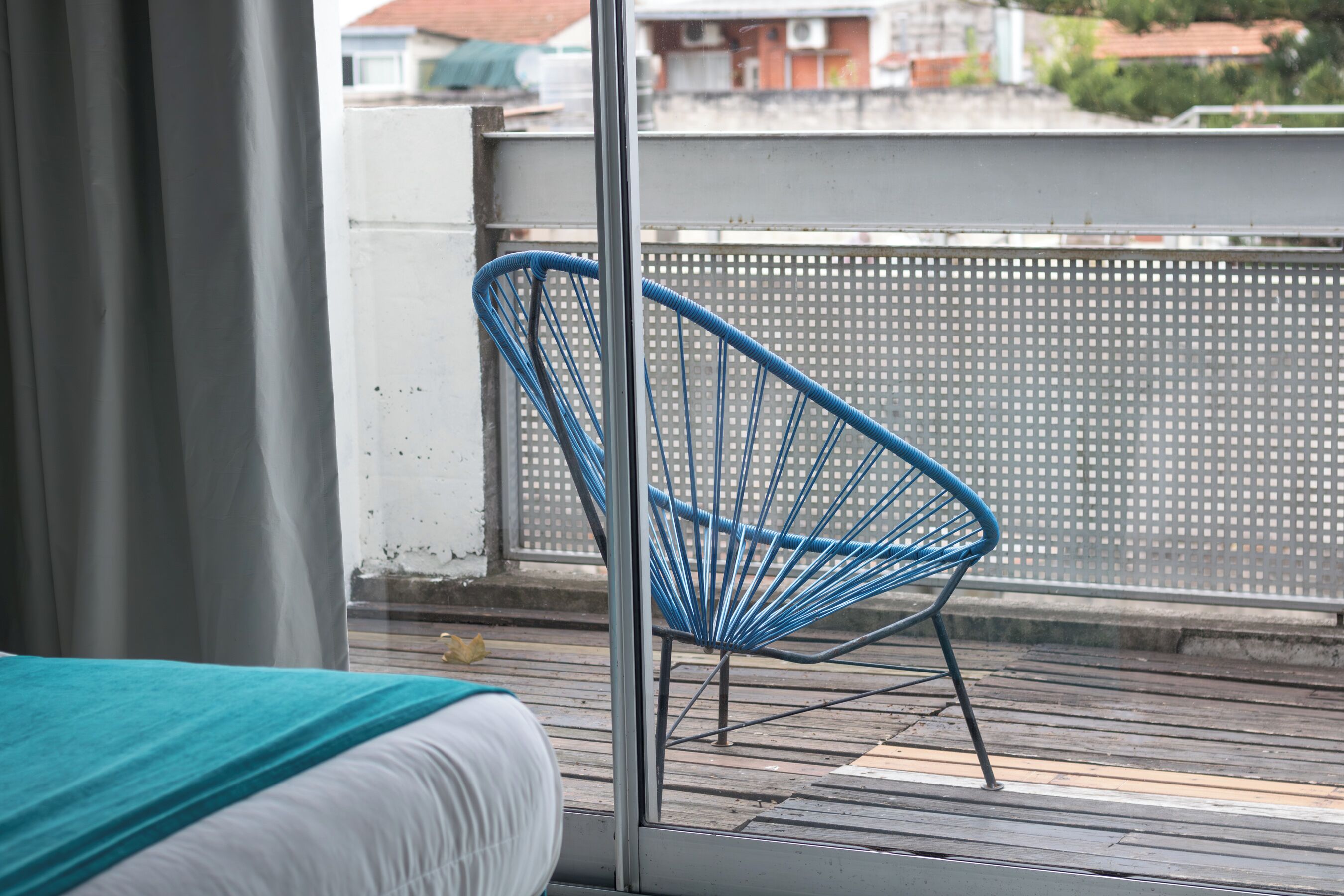 A balcony with a blue chair, a bed with a white and teal bedspread, and a view of buildings in the background.
