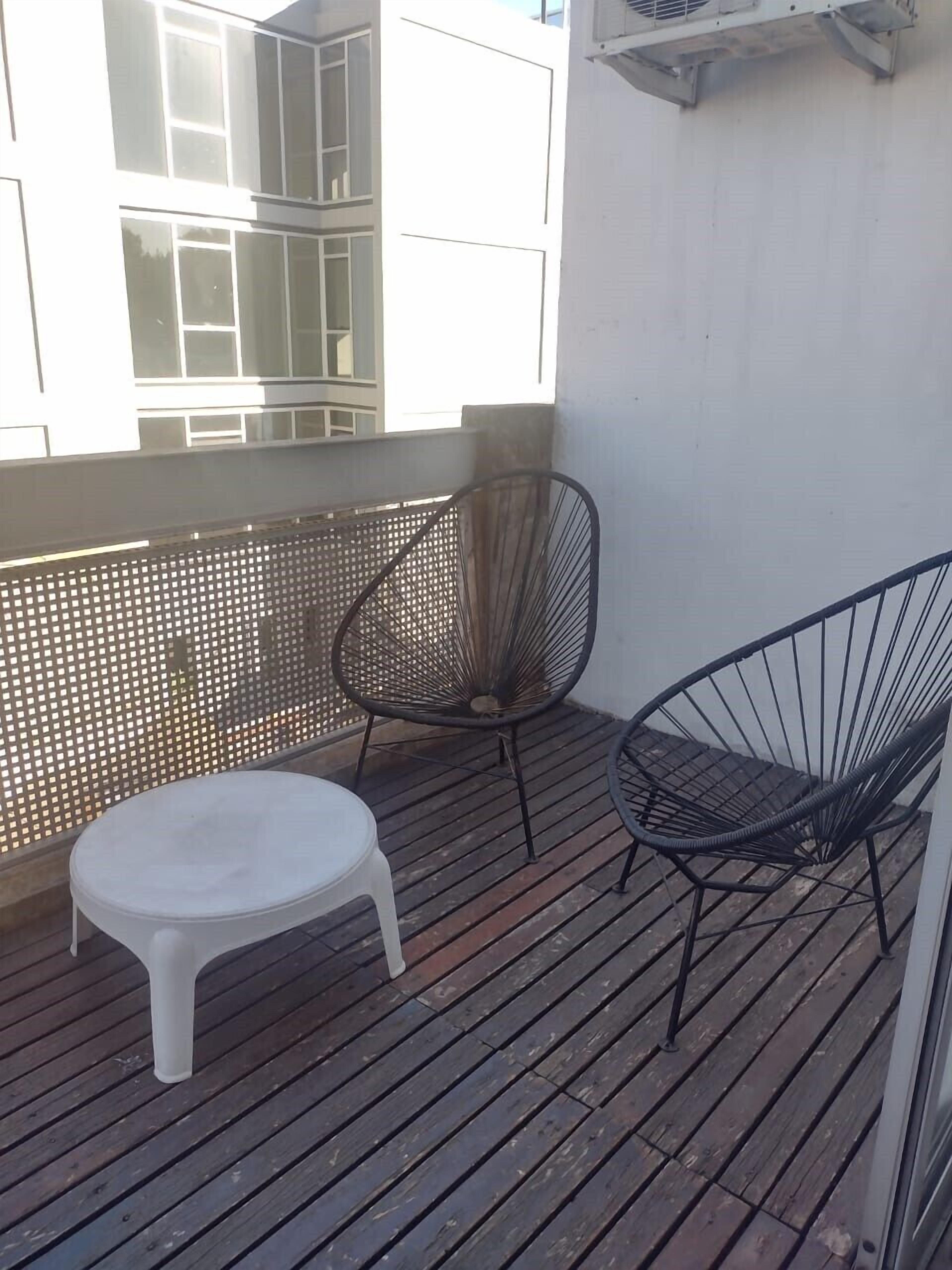 Een balkon met een witte plastic kruk, twee metalen stoelen en een houten vloer.