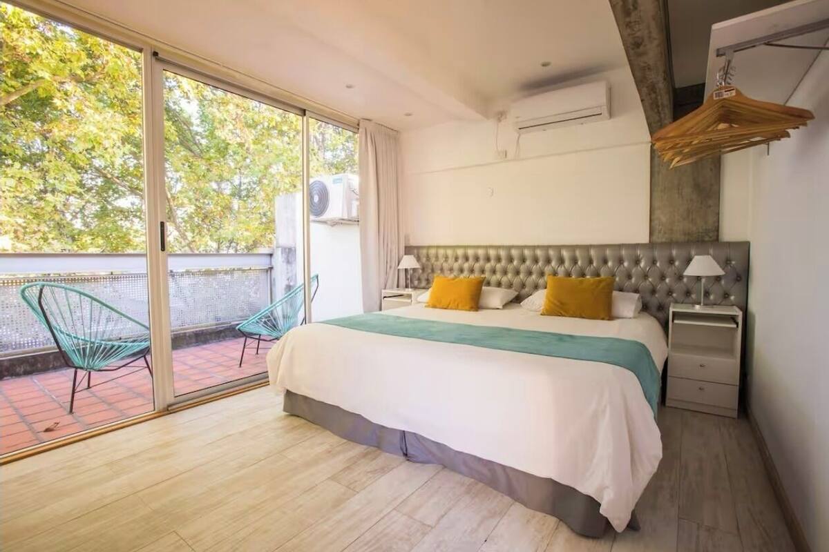 A bedroom with a large bed, two bedside tables, a hanging light fixture, and a sliding glass door leading to an outdoor area with chairs.
