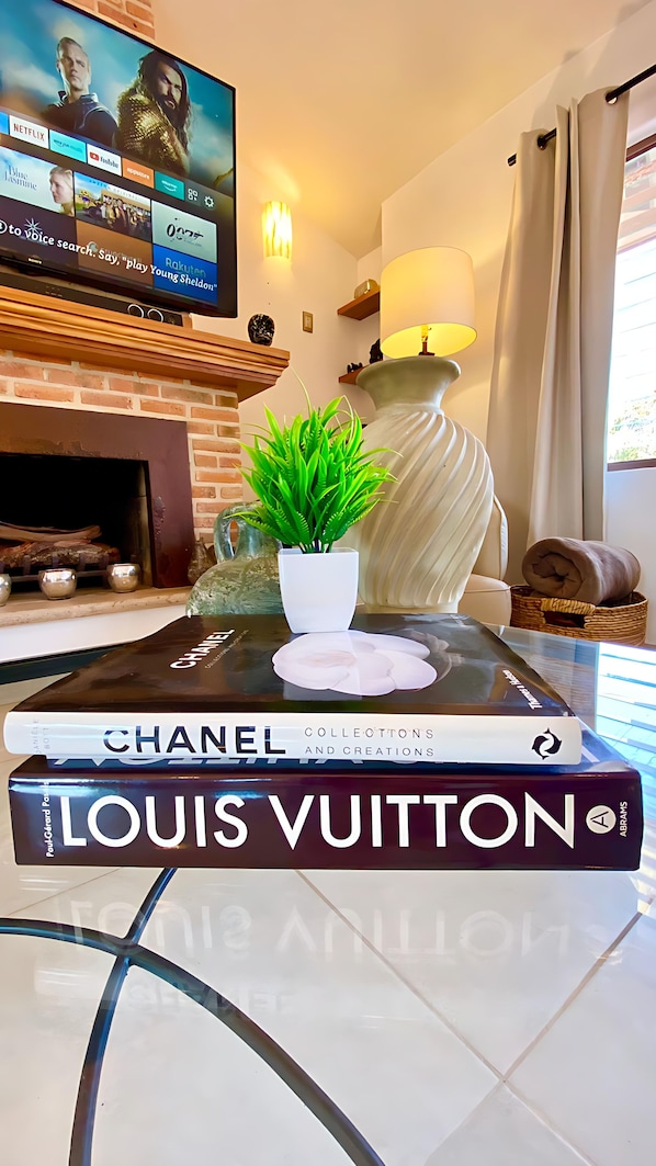 A living room with a TV displaying a movie, a fireplace, a lamp, a potted plant, and a stack of books including 'Chanel' and 'Louis Vuitton'.