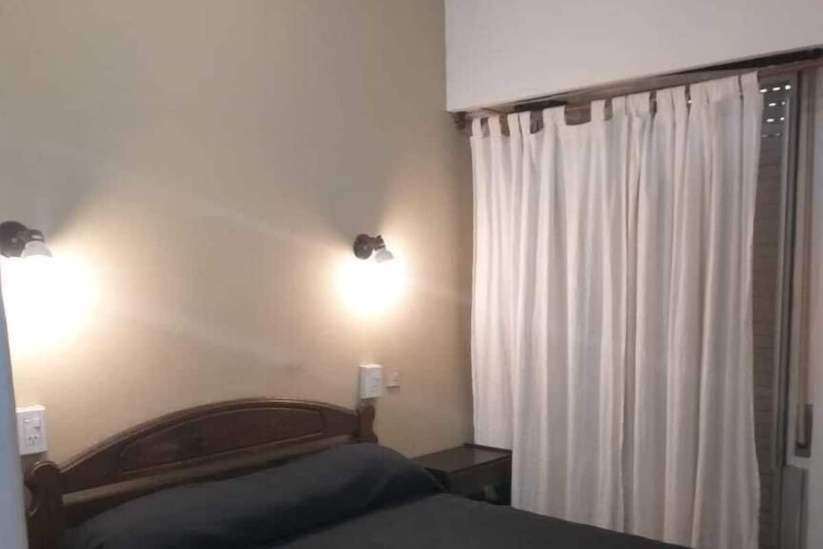 Une chambre à coucher avec un lit en bois, deux lampes fixées au mur et une fenêtre avec des rideaux.