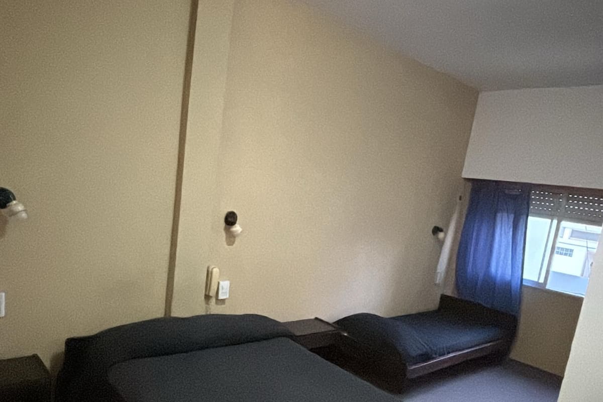 Une chambre d’hôtel avec deux lits, une petite fenêtre avec des rideaux, un luminaire au plafond et un thermostat fixé au mur.