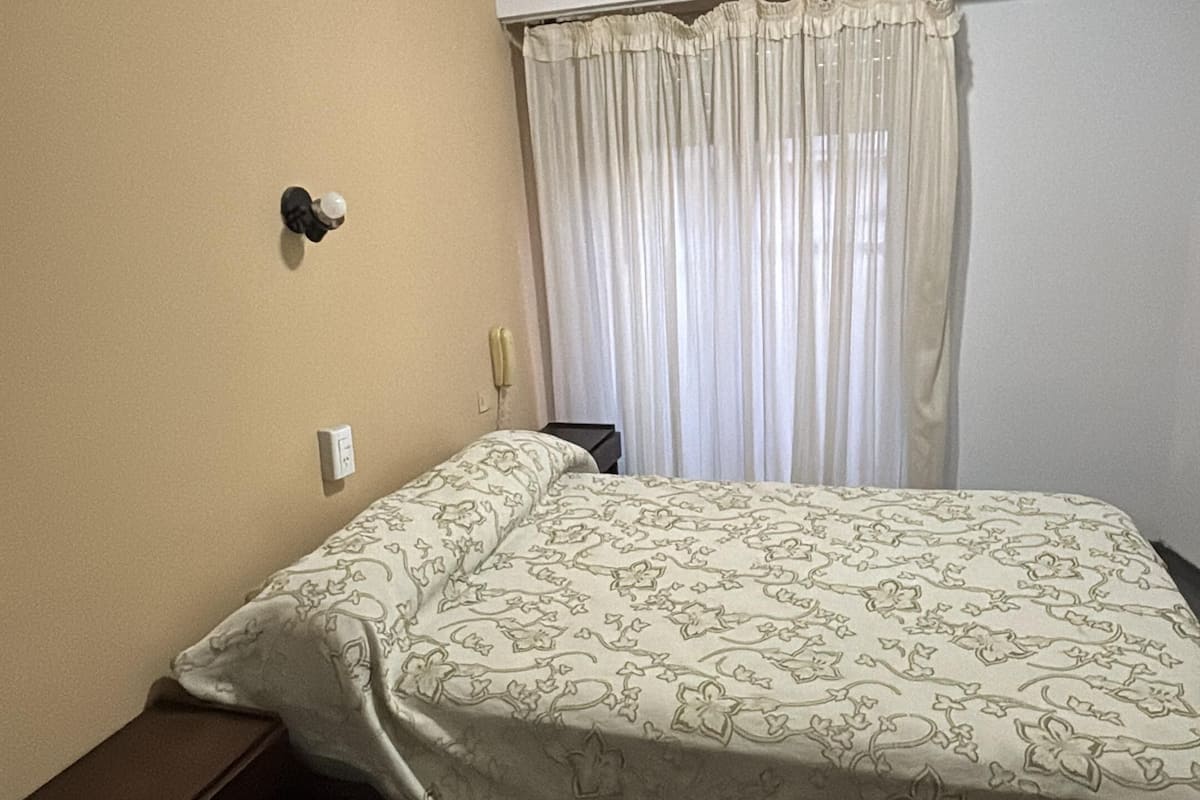 Une chambre d’hôtel avec un lit, une table de chevet, un ventilateur de plafond et une fenêtre avec des rideaux.