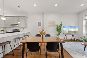 Eine moderne Küche mit weißen Schränken, einem Holzespeisetisch und schwarzen Stühlen. An der Wand hängt ein Gemälde und auf dem Boden steht eine Pflanze.