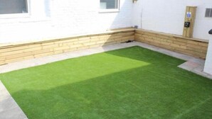 Un petit patio clos, doté d’une pelouse artificielle, de bordures en bois et d’un mur blanc percé d’une fenêtre et d’une porte.