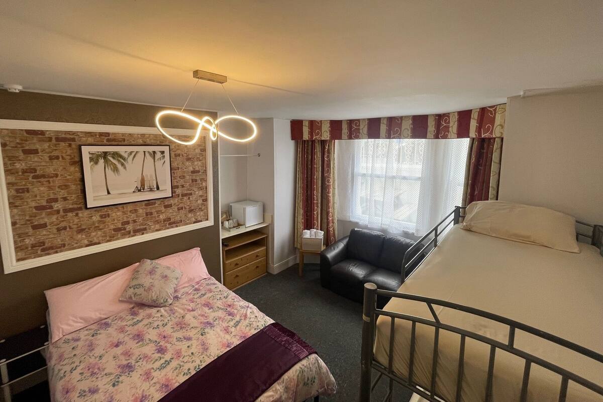 Une chambre d’hôtel avec un lit, un bureau, une chaise, une petite table, une bibliothèque, un tableau au mur et une fenêtre avec des rideaux.