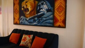 A room with a dark blue sofa and two orange pillows, a large abstract painting on the wall, and a partial view of a patterned wall. - BackpackerKilla (Machu Picchu)