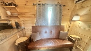 L'intérieur d'un chalet accueillant, avec un plancher en bois, un canapé avec des coussins, une petite table en bois et un coin cuisine doté d'un comptoir et d'ustensiles.