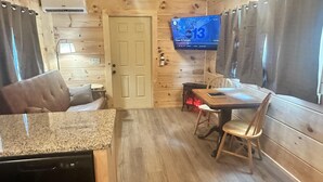 A cozy cabin interior with a wooden interior, a dining area with a table and chairs, a television mounted on the wall, and a door leading to another room. - Hideaway Holler - Hot Tub, Fireplace, Wi-Fi (Union Mills)