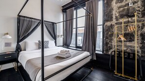 Una cama con dosel de estructura negra, ropa de cama blanca y una manta gris. A la derecha, se ve un perchero dorado.