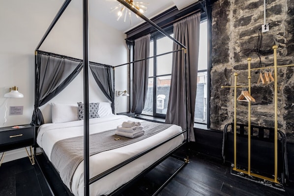 A four-poster bed with a black frame, white bedding, and a grey throw. A gold coat rack is visible to the right.
