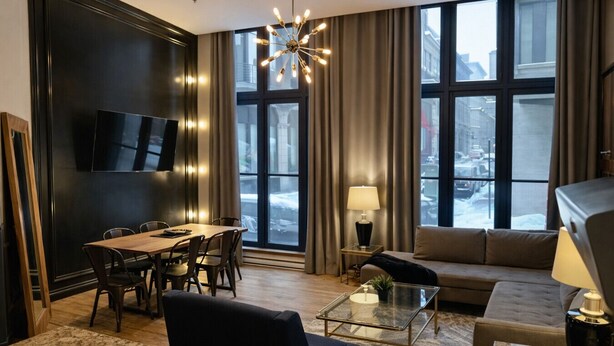 A modern hotel room with a dining area, a sofa, a glass coffee table, and a TV mounted on the wall.