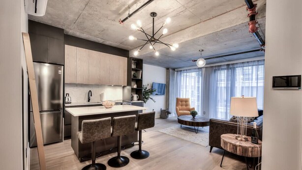 Una cocina moderna con una isla central, taburetes de bar y una mesa de comedor. Hay un sofá y una lámpara en la sala de estar.