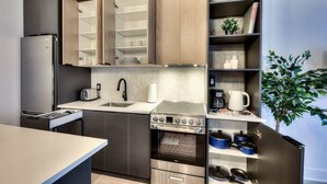Cocina compacta con horno integrado, microondas y refrigerador. Cuenta con armarios de madera y una repisa con utensilios de cocina.