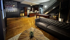 Un vaste espace de vie comprenant un canapé de couleur sombre, une table basse en bois et une table de billard. On aperçoit à l’arrière-plan un coin cuisine avec des meubles en bois et un plan de travail.