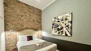Une chambre avec un mur en briques, un lit avec du linge de lit blanc, une tête de lit, une couverture grise, un oreiller et un tableau accroché au mur.