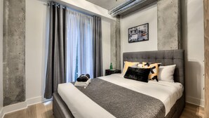 Une chambre moderne avec un grand lit, une literie aux tons de gris et de blanc, une tête de lit grise et une fenêtre agrémentée de rideaux gris.
