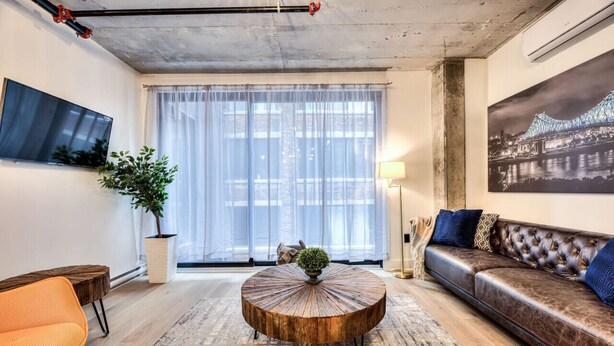 Een moderne woonkamer met een ronde salontafel, een leren bank, een aan de muur bevestigde televisie en een groot raam met dunne gordijnen.