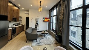 A modern kitchen with wooden cabinets, a built-in microwave, and a stainless steel refrigerator. There is a gray sofa and a small round table with a plant. A large window offers a view of the city. - Luxury 2BD Loft-Old Montreal-2B (Montréal)
