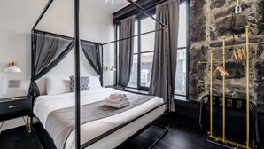 A four-poster bed with a black frame and grey curtains, a wooden floor, a black nightstand with a lamp, and a stone wall. - Luxury 2BD Loft-Old Montreal-2B (Montréal)