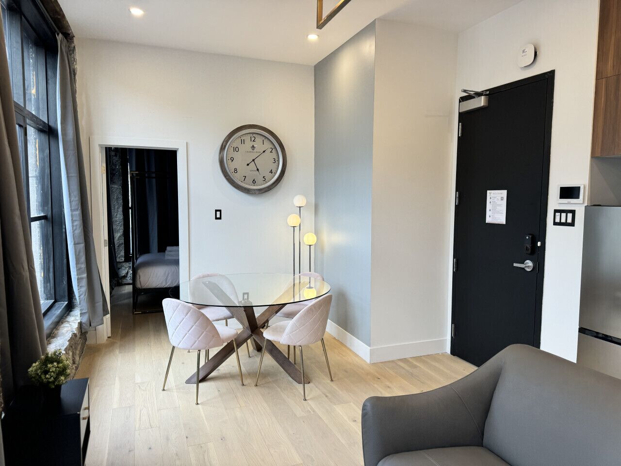 A modern living room with a glass dining table, two chairs, a sofa, a black door, and a wall clock.
