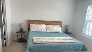 Een slaapkamer met een houten bedframe, blauwgroene beddengoed, twee bedlampen en een raam met zonwering.