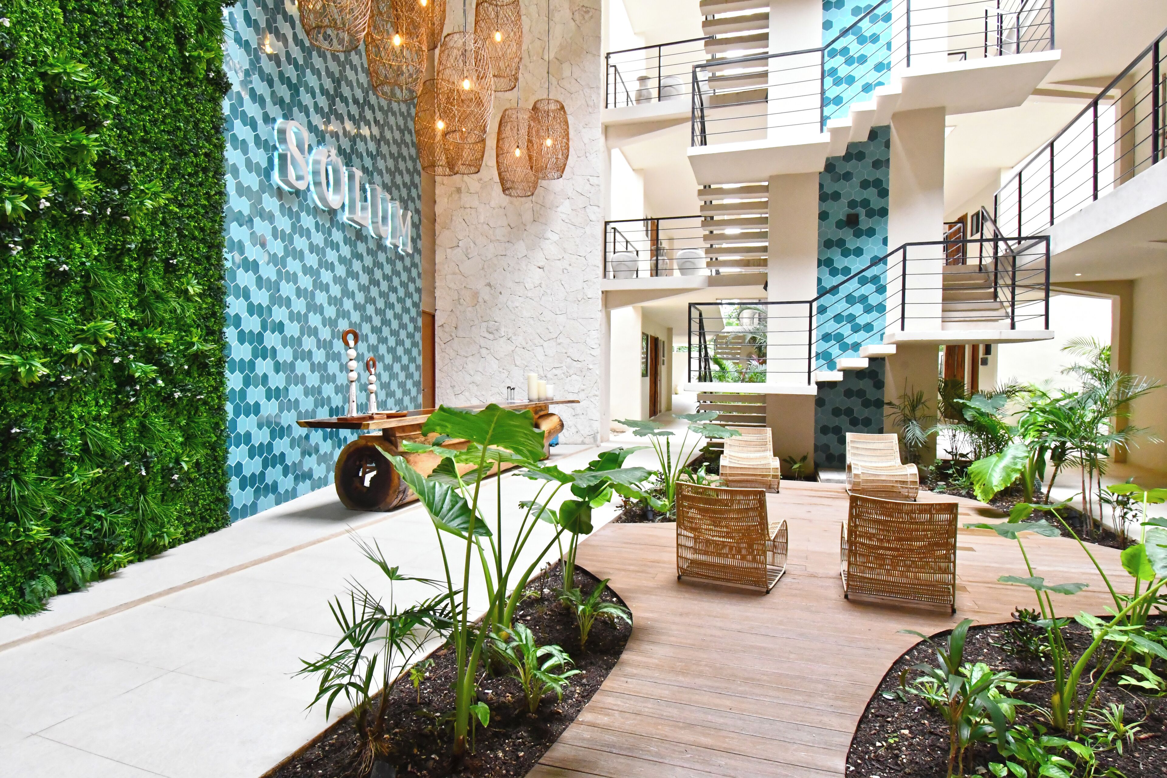 A modern lobby with a green wall, a central planter with plants, wicker furniture, and a staircase.
