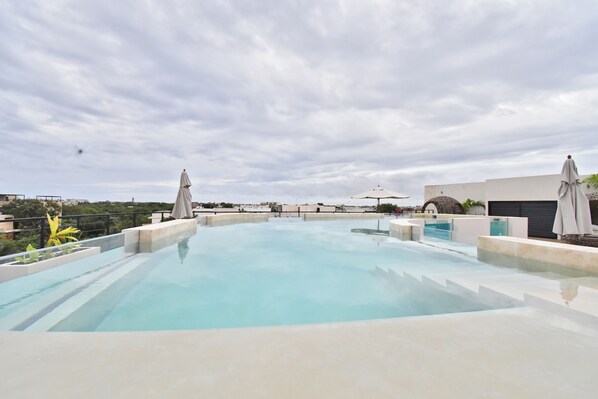 A rooftop swimming pool with clear blue water, surrounded by lounge chairs and umbrellas, and a view of the cityscape in the background. - Solum Living by The Spot Rentals (Tulum)