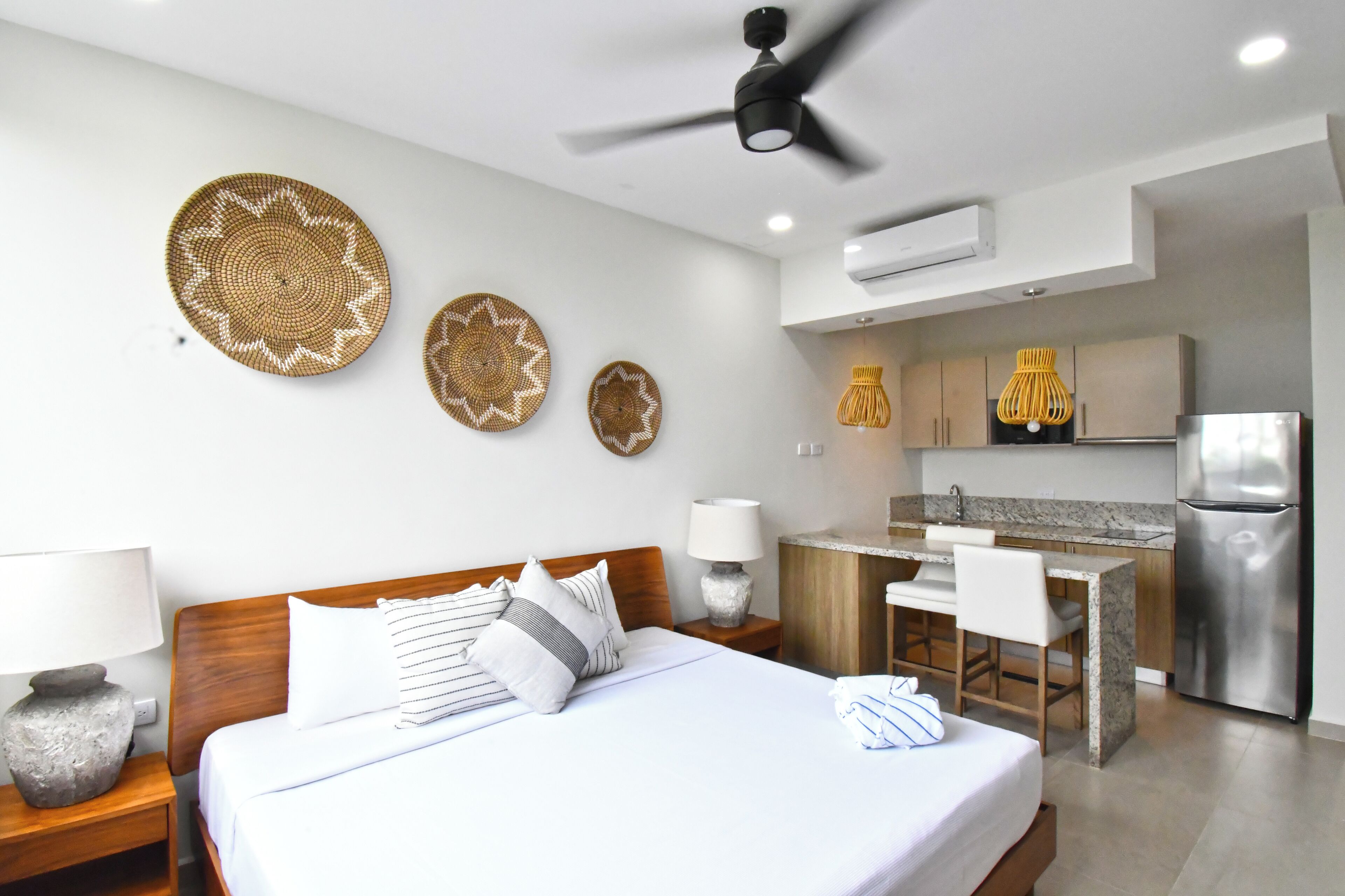 A modern hotel room with a bed, bedside tables, a desk, and a kitchenette.