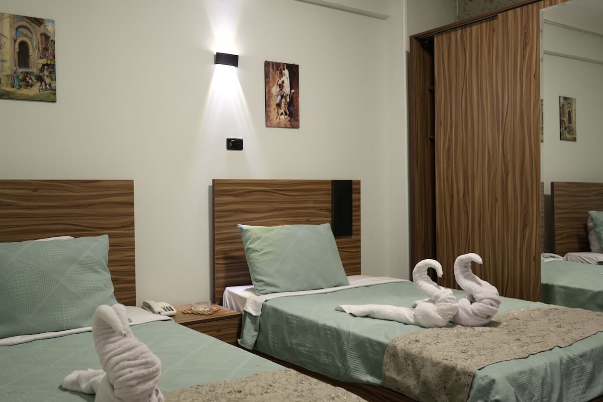 A hotel room with two beds, each with a wooden headboard, light green bedding, and matching pillows. There are wall-mounted light fixtures and artwork above the beds.