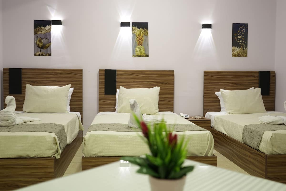 A hotel room with three beds, each with a wooden headboard, white bedding, and a towel sculpture. There are wall-mounted light fixtures and artwork above the beds.