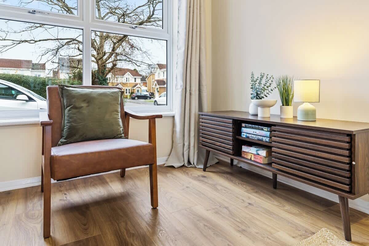 A room with a wooden chair, a dark wooden side table with books and a lamp, and a window with a view of houses and a car outside.