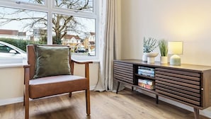 Une pièce avec une chaise en bois, une petite table en bois foncé sur laquelle se trouvent des livres et une lampe, et une fenêtre donnant sur des maisons et une voiture à l’extérieur.