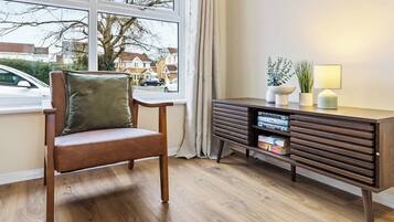 A room with a wooden chair, a dark wooden side table with books and a lamp, and a window with a view of houses and a car outside.