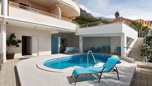 A modern building with a swimming pool, outdoor seating, and a mountain view. - Apartments Relax Brela - Two Bedroom Apartment (Brela)