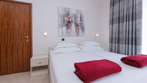 A hotel room with a bed, a wooden door, a white nightstand, a red towel, a gray curtain, and a painting on the wall. - Apartments Relax Brela - Studio (Brela)