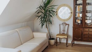 A room with a white sofa, a wooden chair, a potted plant, a round mirror, and a wooden cabinet with glass doors. - Enchanted Misty Valley Cottage Apartments - Standard One Bedroom Apartment (Sentjost nad Horjulom)