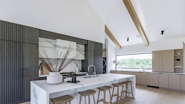 A modern kitchen with a marble island, wooden stools, and a large window providing a view of greenery.