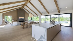 A spacious kitchen with a marble island, wooden beams, and large windows offering a view of greenery.