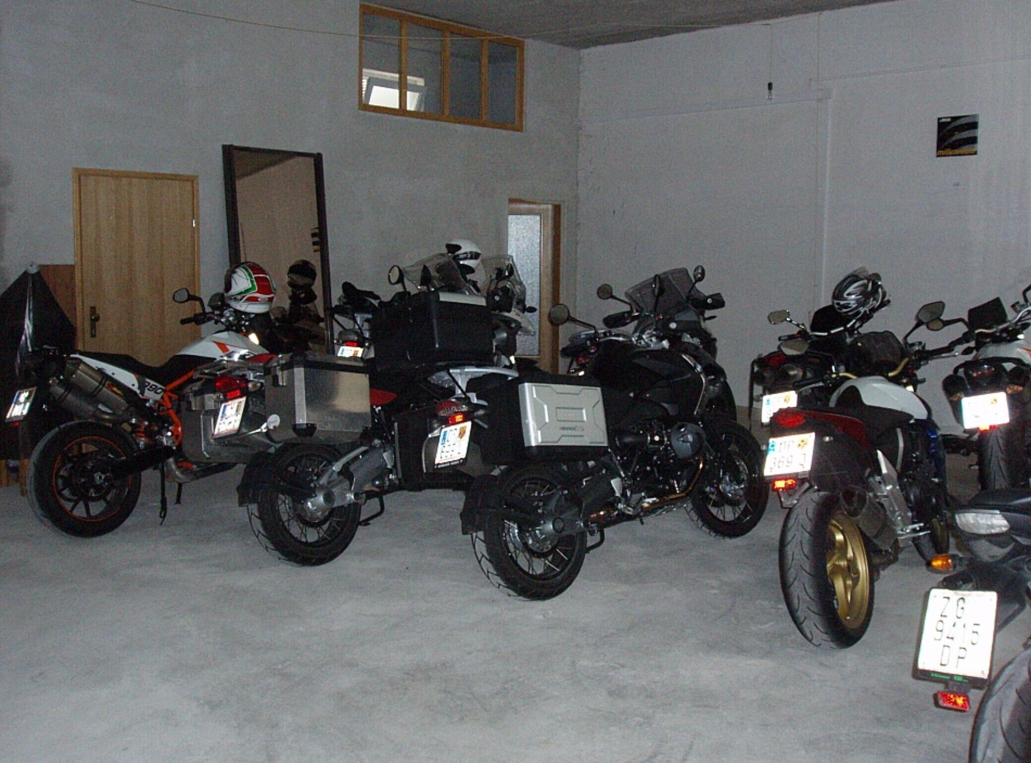 A row of motorcycles parked indoors with a door and a window visible in the background.