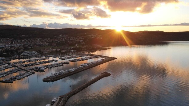 An aerial view of a marina with boats docked, a dock extending into the water, and a cityscape in the background during sunset.