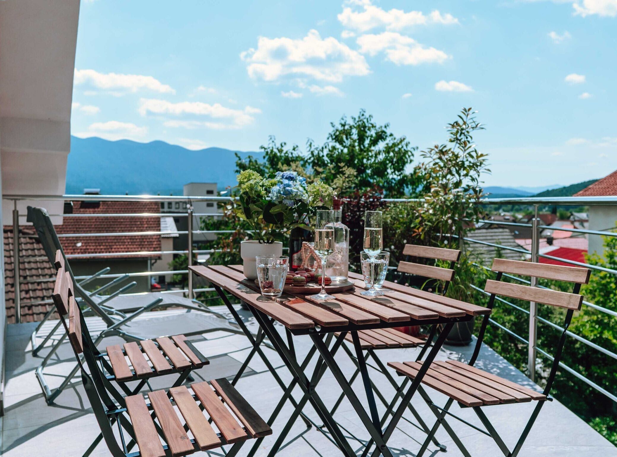 Eine Terrasse im Freien mit einem Holztisch und -stühlen, einer Blumenvase und Blick auf die Berge.